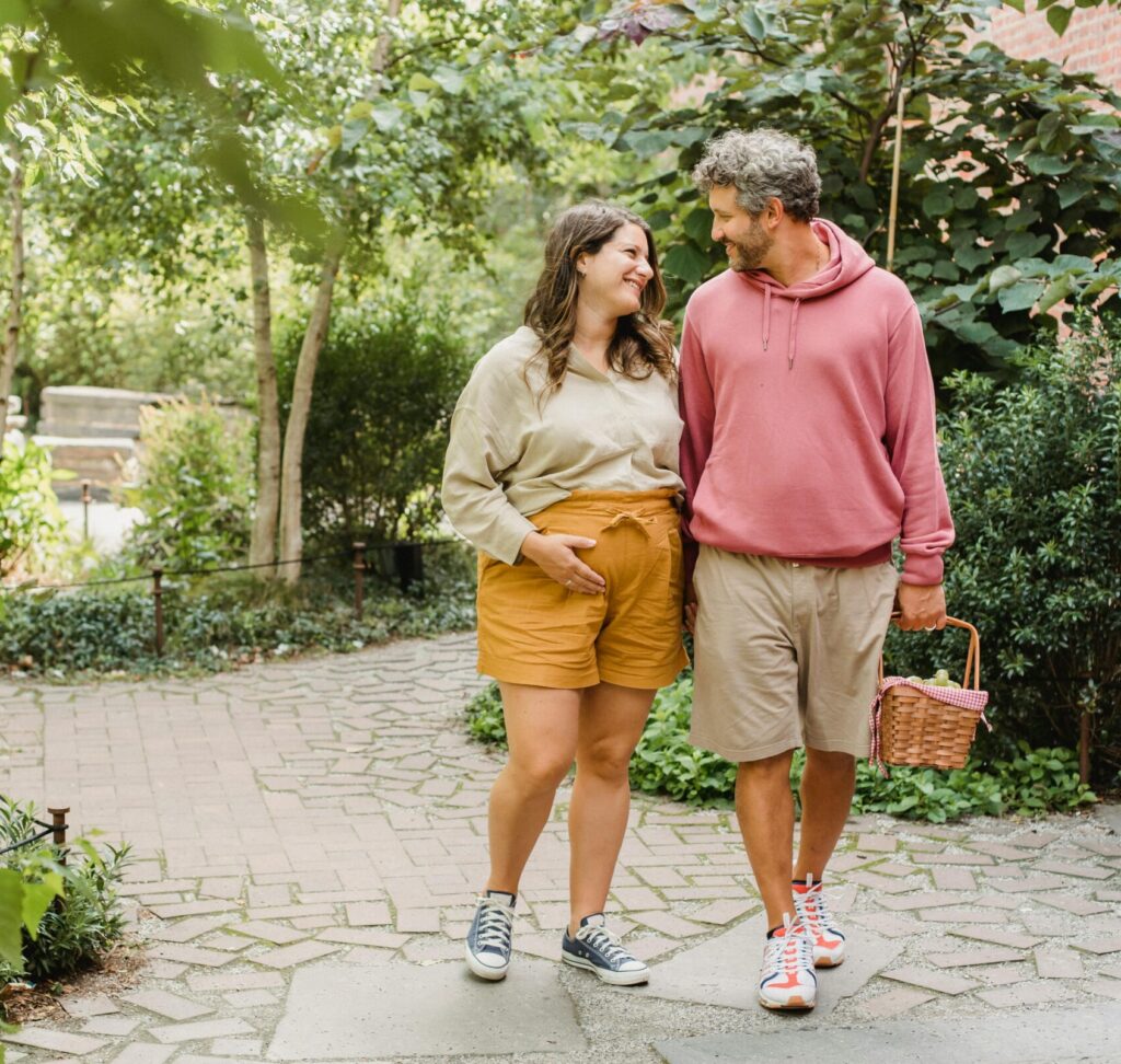 pregnant couple walking outside in the center of a path while smiling at each other