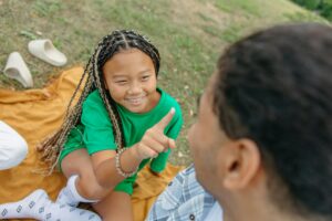 child with pulled up socks sits on a picnic blanket on the lawn and smiles up at her parent